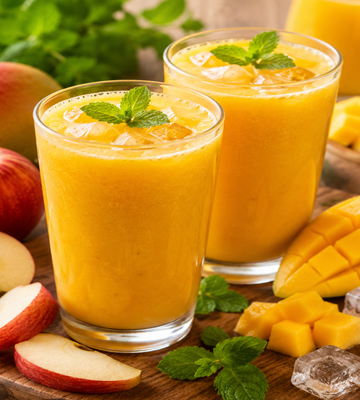 Twee glazen mango appel sap met ijsblokjes, verse mango, appelpartjes en muntblaadjes op een houten plank