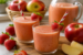 Twee glazen romig appel aardbei sap met verse aardbeien als garnering, geserveerd op een houten plank met appels, aardbeien en een karaf sap op de achtergrond