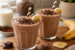 Twee romige banaan chocolade milkshakes in glazen, gegarneerd met chocoladeschaafsel en banaanschijfjes, geserveerd op een houten plank met bananen en chocolade op de achtergrond