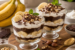 Twee glazen desserts met lagen room, banaan en chocolade, gegarneerd met banaanschijfjes, chocoladeschaafsel en munt, geserveerd op een houten plank