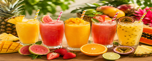 Een rij glazen met verschillende zoete fruitsappen in diverse kleuren, omringd door vers tropisch fruit zoals aardbeien, sinaasappel, ananas en passievrucht.