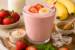 Romige aardbei proteïne milkshake in een glas, gegarneerd met aardbeien en banaanschijfjes. Op de achtergrond staan een fles melk, verse aardbeien, bananen, ijsblokjes, honing en een schaaltje eiwitpoeder.