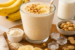 Romige banaan-pindakaas proteïne milkshake in een glas, afgewerkt met banaanschijfjes en stukjes noot. Op de achtergrond staan bananen, melk, pinda’s, ijsblokjes en een schaaltje eiwitpoeder.