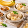 Kommen met romig bananenijs, gegarneerd met bananenschijfjes, gehakte noten en verse munt, geserveerd naast honing en verse bananen.