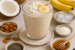 Romige kokos-chia milkshake met banaan als garnering, omringd door kokos, amandelen, chiazaad, honing en ijsblokjes.