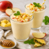 Twee romige sinaasappel-appel milkshakes in glazen met stukjes appel en munt als garnering, omringd door appelpartjes, ananasstukjes, honing en ijsblokjes op een lichte ondergrond.